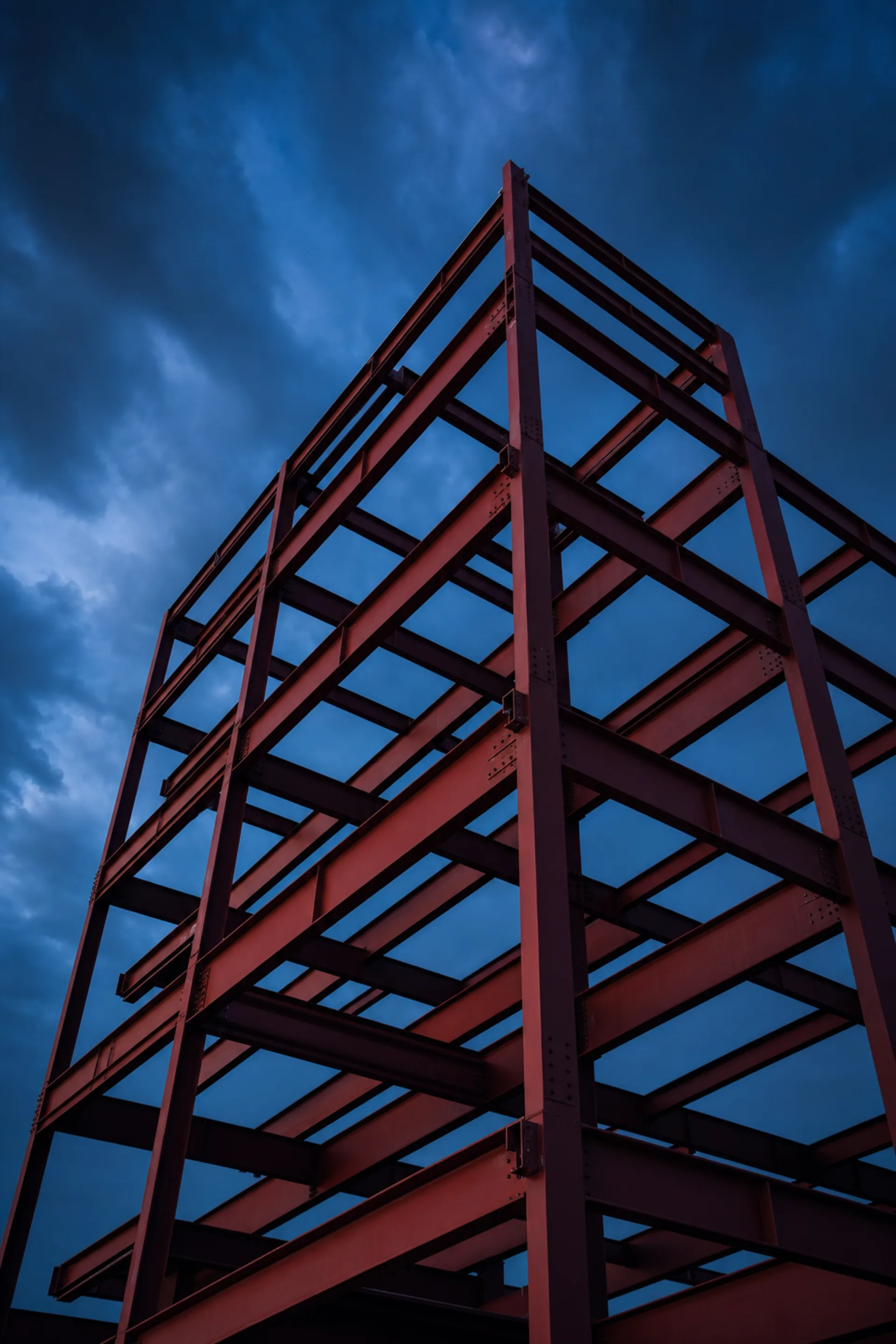 Exposed red structural steel frame of a building under construction against a twilight sky